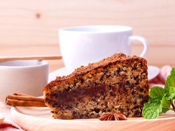 ein Stück Kuchen auf einem Holzteller, dahinter eine weiße Tasse auf Unterteller