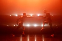 Zwei Tänzer in dynamischer Pose auf einer Bühne mit intensiver roter Beleuchtung und Lichtquellen im Hintergrund