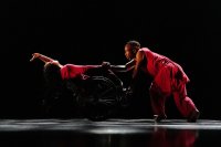 Zwei Tänzer in roten Outfits, einer im Rollstuhl, der andere hält dessen Hand und lehnt sich nach vorne auf einer dunklen Bühne.