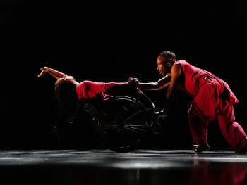 Foto: UnmuteDanceTheatre
Zwei Tänzer in roten Outfits, einer im Rollstuhl, der andere hält dessen Hand und lehnt sich nach vorne auf einer dunklen Bühne.