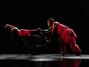Zwei Tänzer in roten Outfits, einer im Rollstuhl, der andere hält dessen Hand und lehnt sich nach vorne auf einer dunklen Bühne.