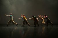 Fünf Tänzer in farbigen Oberteilen und braunen Hosen bewegen sich synchron mit ausgestreckten Armen auf einer dunklen Bühne