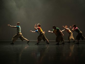Fünf Tänzer in farbigen Oberteilen und braunen Hosen bewegen sich synchron mit ausgestreckten Armen auf einer dunklen Bühne