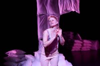 Person in heller Kleidung vor einem Stapel Kissen und einem hängenden Stoffstück in dunkler Umgebung