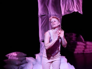 Foto: Ludwig Olah
Person in heller Kleidung vor einem Stapel Kissen und einem hängenden Stoffstück in dunkler Umgebung