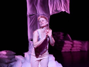 Person in heller Kleidung vor einem Stapel Kissen und einem hängenden Stoffstück in dunkler Umgebung