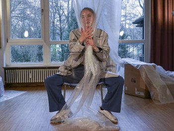 Person sitzt breitbeinig auf einem Stuhl in einem Raum mit Holzboden und hält eine große Luftpolsterfolie