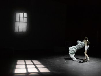 Foto: Quinn Maier
Person in Unterwäsche wirbelt ein großes, weißes, textilähnliches Objekt auf einer dunklen Bühne mit Fensterlichtreflexionen
