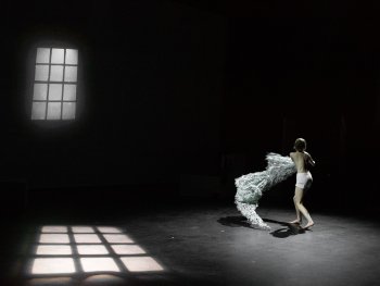 Person in Unterwäsche wirbelt ein großes, weißes, textilähnliches Objekt auf einer dunklen Bühne mit Fensterlichtreflexionen