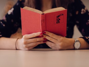 Eine Frau mit rotem Nagellack hält ein rotes Buch aufgeschlagen in Händen