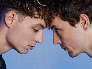 Zwei junge Männer mit dunklen Haaren und Halsketten stehen sich frontal gegenüber vor blauem Hintergrund