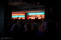 Dunkler Innenraum mit einer Bar im Hintergrund, an der mehrere Personen stehen, davor eine Gruppe sitzender Zuschauer.