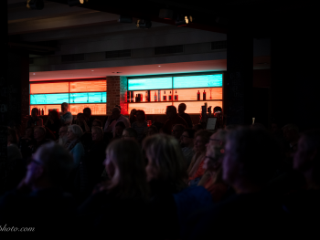 Dunkler Innenraum mit einer Bar im Hintergrund, an der mehrere Personen stehen, davor eine Gruppe sitzender Zuschauer.