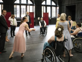 Gruppe von Menschen in einem großen Raum, einige im Rollstuhl, andere in eleganter Kleidung, die sich in einem Kreis versammeln