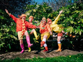 Die 4 Schauspielerinnen in bunter Kleidung und und bunter Gummistiefel stehen auf einem Bein
