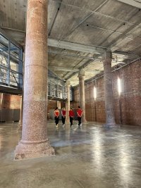 Vier Tänzer*innen in Rückenansicht tanzen nebeneinander, zwischen zwei großen Säulen aus Granit in einem großen Saal mit Steinmauern.