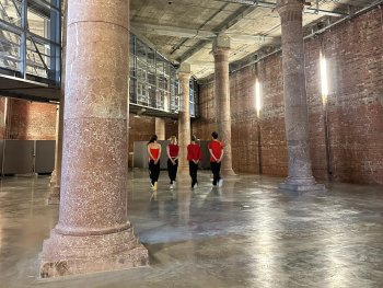 Copyright: Astrid Betz Vier Tänzer*innen in Rückenansicht tanzen nebeneinander, zwischen zwei großen Säulen aus Granit in einem großen Saal mit Steinmauern.
