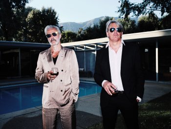 Zwei Männer in Anzügen stehen nebeneinander an einem Pool, jeder hält ein Glas in der Hand.