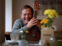 Person hält Ukulele vor sich sitzend an einem Tisch mit mehreren Blumenvasen und Tassen