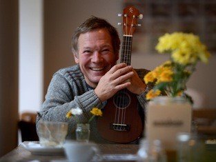 Person hält Ukulele vor sich sitzend an einem Tisch mit mehreren Blumenvasen und Tassen