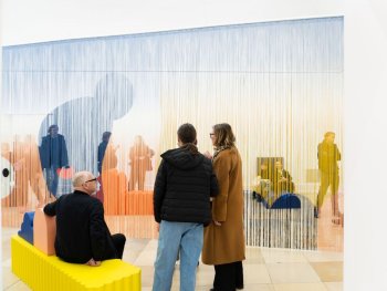 Copyright: Erika Hock, Yellow, 2023 & Curtain (straight/blue figure), 2020; Courtesy Erika Hock & COSAR, Düsseldorf Gelbes Sitzmöbel im Vordergrund, dahinter ein farbiger Vorhang aus Fäden von Erika Hock