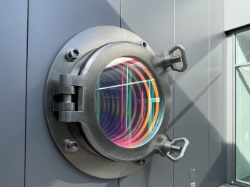 Copyright: Sebastian Kuhn, Bullauge, 2024 Edelstahl, Licht, Glas, Spiegel, B 210 x H 210 x T 65 cm Ein geschlossenes Bullauge in dessen Innerem sich das Licht in den Farben Blau, Orange und Rot bricht.