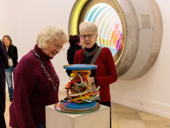 Copyright: Sebastian Kuhn Beauty Trophy LV, 2025 // Arrival, 2026; (c) VG Bild-Kunst Bonn, 2026 Ein moderner Pokal aus Metallscheiben und einer Stange, um die bunte Kunststoffstreifen lose gewickelt sind, im Hintergrund ein verspiegeltes und beleuchtetes Airbus-Triebwerk von Sebastian Kuhn