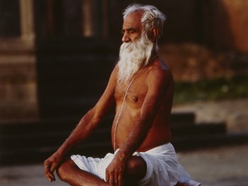 Mann mit langem, weißen Bart und nacktem Oberkörper im Yogasitz an einer Straße in Indien