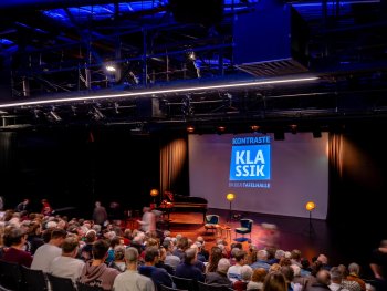 Bühne mit Klavier, zwei Stühlen und Leinwand mit Schriftzug 'KONTRASTE KLASSIK IN DER TAFELHALLE' vor Publikum in einem Theaterraum