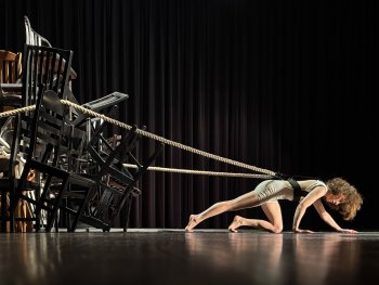 Foto: Wolfgang Keller
Frau in Sportkleidung in einer Plank-ähnlichen Pose auf einer Bühne, im Hintergrund mehrere übereinander gestapelte Stühle, verbunden mit einem dicken Seil.