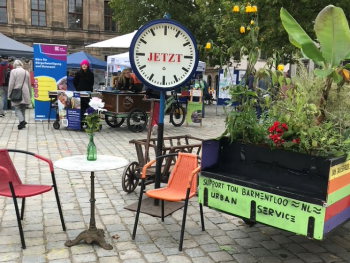 Große Standuhr mit der Aufschrift „Jetzt“ steht zwischen Stühlen, einem kleinen Tisch mit Blumenvase und einem bepflanzten Wagen auf einem Marktplatz mit Infoständen im Hintergrund.