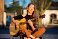 Frau mit langen Haaren sitzt im Schneidersitz auf dem Boden und spielt akustische Gitarre im Freien