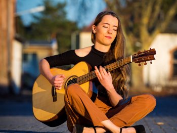 Foto: Reinhold Blaha
Frau mit langen Haaren sitzt im Schneidersitz auf dem Boden und spielt akustische Gitarre im Freien