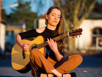 Frau mit langen Haaren sitzt im Schneidersitz auf dem Boden und spielt akustische Gitarre im Freien