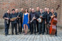 Das Foto zeigt die zwölfköpfige Band in schwarz gekleidet und mit ihren Instrumenten vor einer roten Backsteinmauer. In der Mitte steht die Sängerin der Band in einem blauen Kleid.
