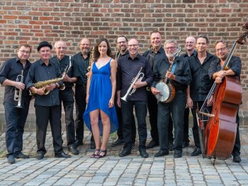Das Foto zeigt die zwölfköpfige Band in schwarz gekleidet und mit ihren Instrumenten vor einer roten Backsteinmauer. In der Mitte steht die Sängerin der Band in einem blauen Kleid.