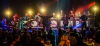 Das Foto zeigt einen abendlichen Bühnenauftritt der Band im Kulturgarten des Künstlerhauses. Unten sitzen die Gäste auf Bierbänken, auf der Bühne singen und spielen die Bandmitglieder voller Energie und bunt beleuchtet.