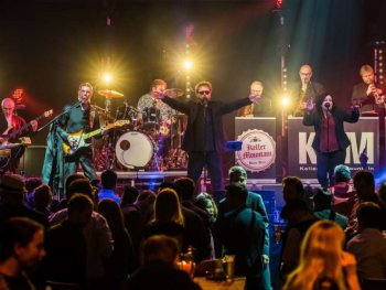 Das Foto zeigt einen abendlichen Bühnenauftritt der Band im Kulturgarten des Künstlerhauses. Unten sitzen die Gäste auf Bierbänken, auf der Bühne singen und spielen die Bandmitglieder voller Energie und bunt beleuchtet.