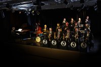 Das Foto zeigt das Groove Legend Orchestra bei einem stimmungsvoll beleuchteten Bühnenauftritt. Rechts sitzen die Bandmitglieder mit den Blech- und Blasinstrumenten, links sitzen der Schlagzeuger, der Kontrabassspieler und mit dem Rücken zum Publikum, der Pianist.
