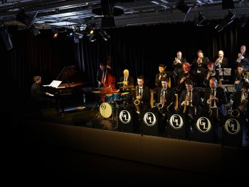 Foto: Marc Pfeifer
Das Foto zeigt das Groove Legend Orchestra bei einem stimmungsvoll beleuchteten Bühnenauftritt. Rechts sitzen die Bandmitglieder mit den Blech- und Blasinstrumenten, links sitzen der Schlagzeuger, der Kontrabassspieler und mit dem Rücken zum Publikum, der Pianist.