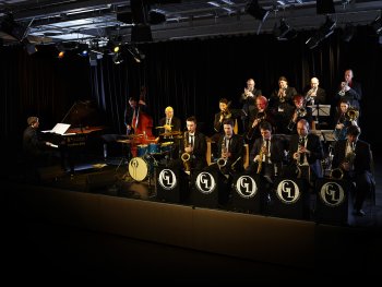 Das Foto zeigt das Groove Legend Orchestra bei einem stimmungsvoll beleuchteten Bühnenauftritt. Rechts sitzen die Bandmitglieder mit den Blech- und Blasinstrumenten, links sitzen der Schlagzeuger, der Kontrabassspieler und mit dem Rücken zum Publikum, der Pianist.