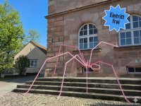 Große, rosa Metallskulptur einer Spinne mit langen, dünnen Beinen vor einem Sandsteingebäude
