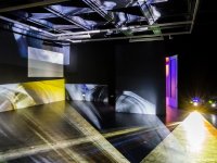 Dunkler Raum mit mehreren projizierten Licht- und Bildflächen sowie offenstehender Tür mit violettem Lichtschein