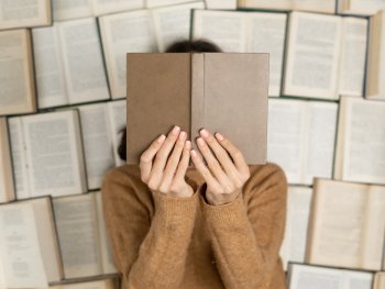 Eine Person in einem ockerfarbenen Pullover hält sich mit beiden Händen ein Buch vor das Gesicht. Die Person liegt auf vielen aufgeschlagenen Büchern.