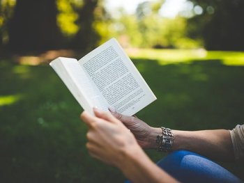 Person hält aufgeschlagenes Buch in beiden Händen im Freien sitzend