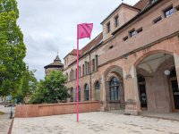 Eingangsbereich eines historischen Sandsteingebäudes mit Arkaden und einem pinkfarbenen Fahnenmast auf gepflastertem Platz