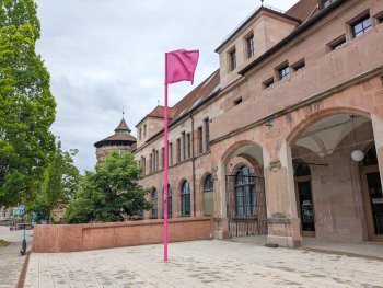 Eingangsbereich eines historischen Sandsteingebäudes mit Arkaden und einem pinkfarbenen Fahnenmast auf gepflastertem Platz