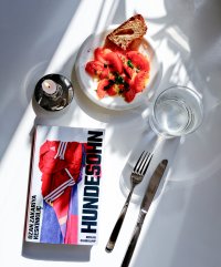 Eingedeckter weißer Tisch mit Schatten, Kerze, Tomatensalat, Wasserglas und dem Roman "Hundesohn"