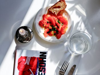 Eingedeckter weißer Tisch mit Schatten, Kerze, Tomatensalat, Wasserglas und dem Roman "Hundesohn"