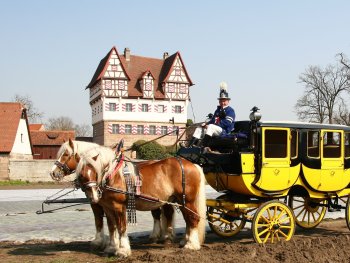 zwei Pferde ziehen einen historische Postkutsche in ländlicher Umgebung.